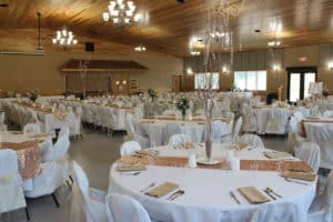 Odessa Event Center decorated with white tablecloths and white chair covers with rose gold accents and centerpieces with tree branches.