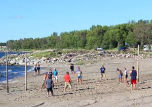 A group of people play a game of sand volleyball with a net next to the lake at Eastbay Campground.