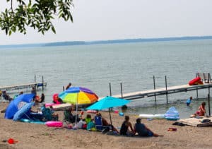 People enjoying a sunny day next to the lake under sun umbrellas on the swimming beach at Eastbay Campground.