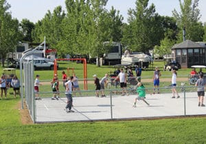 A group of kids, teens, and adults playing a game of basketball on the Eastbay Campground basketball court on a sunny day.