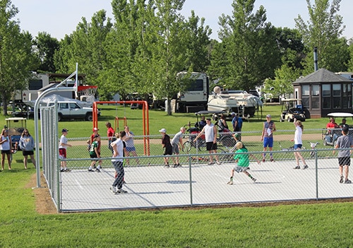 A group of kids, teens, and adults playing a game of basketball on the Eastbay Campground basketball court on a sunny day.