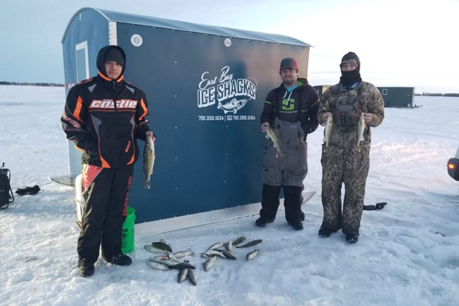 Three bundled up people hold fish that they caught ice fishing while standing outside an Eastbay Campground Ice House rental.